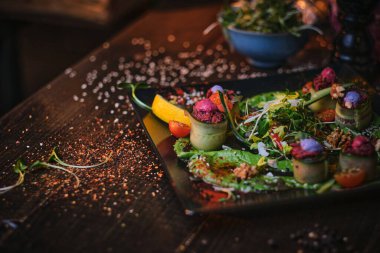 Raw vegetarian plate of appetizer rolls and a bowl of herbs served on a wooden table next to salt grinder in a restaurant