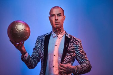Elegant tattooed male model posing for photoshoot in a bright neon studio and holding a golden soccer ball while looking serious