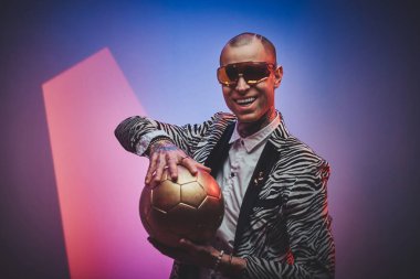 Joyful tattooed male model posing for photoshoot in a neon studio and holding a golden soccer ball while smiling