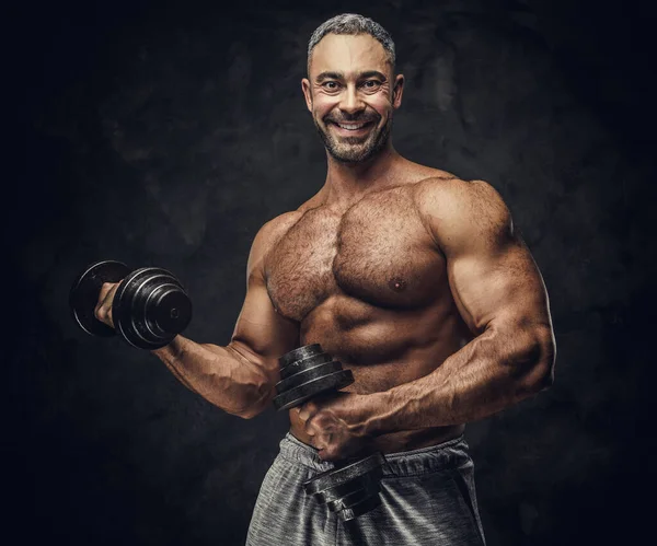 Serious, adult, fit muscular caucasian man coach posing for a photoshoot in a dark studio, wearing sportswear, showing his muscles with dumbbells — Stockfoto