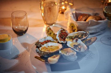 Rich dinner bowl of oysters and bread loafs with sauce served on a table next to candle, a glass of wine, and a wicker basket of bread in a restaurant