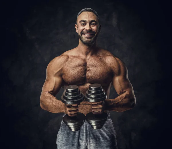Charming, adult, fit muscular caucasian man coach posing for a photoshoot in a dark studio, wearing sportswear, showing his muscles with dumbbells — Stockfoto