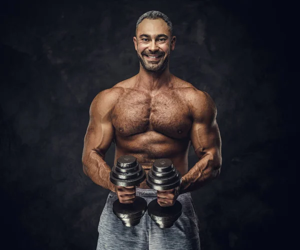 Serious, adult, fit muscular caucasian man coach posing for a photoshoot in a dark studio, wearing sportswear, showing his muscles with dumbbells — 图库照片