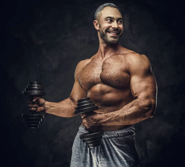 Serious, adult, fit muscular caucasian man coach posing for a photoshoot in a dark studio, wearing sportswear, showing his muscles with dumbbells — Stock Fotó