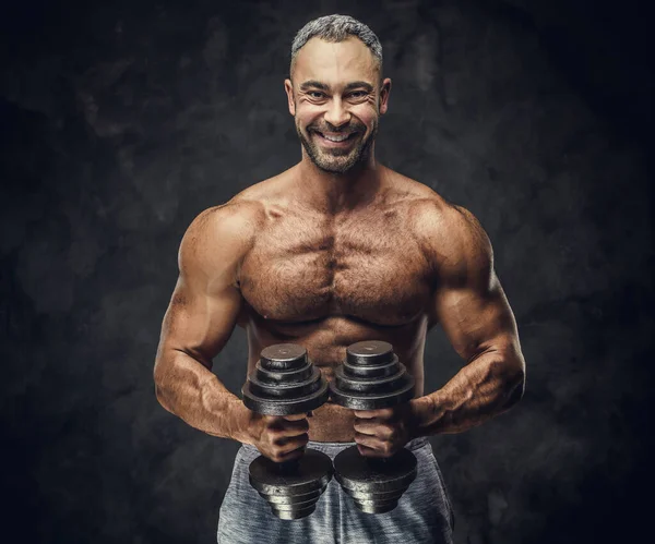 Charming, adult, fit muscular caucasian man coach posing for a photoshoot in a dark studio, wearing sportswear, showing his muscles with dumbbells — 图库照片