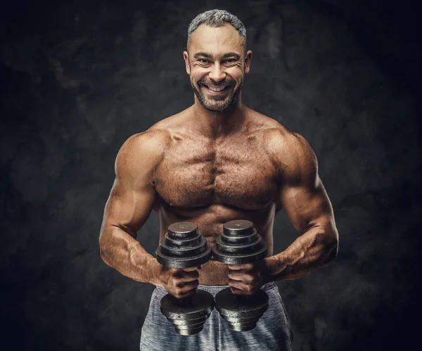 Hombre caucásico encantador, adulto, en forma musculoso entrenador posando para una sesión de fotos en un estudio oscuro, usando ropa deportiva, mostrando sus músculos con mancuernas — Foto de Stock