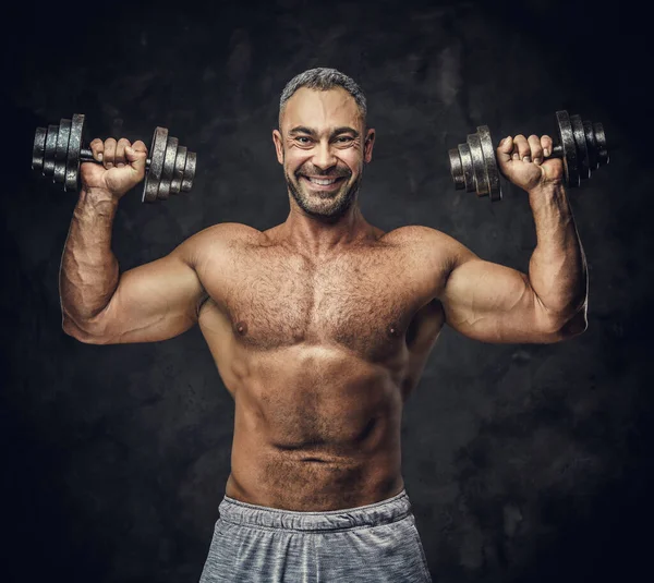 Hombre caucásico encantador, adulto, en forma musculoso entrenador posando para una sesión de fotos en un estudio oscuro, usando ropa deportiva, mostrando sus músculos con mancuernas — Foto de Stock