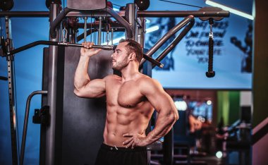 Shirtless bodybuilder posing for a camera while holding a bar, standing next to back pulling machine in a modern fitness club