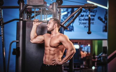 Shirtless bodybuilder posing for a camera while holding a bar, standing next to back pulling machine in a modern fitness club