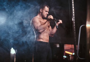 Fit athlete pumping up his biceps on a hand pull machine in a dark gym under the spotlights surrounded by smoke