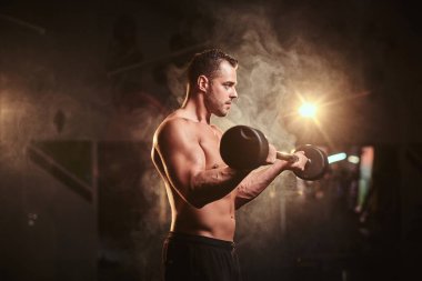 Shirtless bodybuilder doing weightlifts with barbell in a dark gym surrounded by smoke