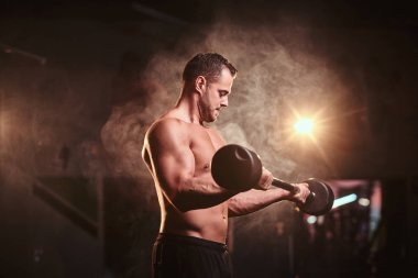 Shirtless bodybuilder doing weightlifts with barbell in a dark gym surrounded by smoke