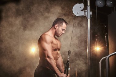 Fit sportsman concentrated on an excersise on a hand pull machine, pumping up his triceps in a dark gym surrounded by smoke