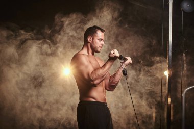 Fit athlete pumping up his biceps on a hand pull machine in a dark gym under the spotlights surrounded by smoke