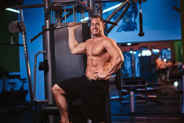 Bodybuilder torse nu posant pour un appareil photo tout en tenant un bar, debout à côté de la machine de traction arrière dans un club de fitness moderne — Photo