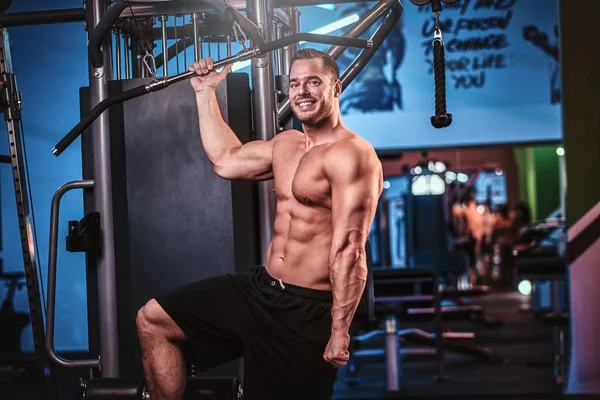 Fisicoculturista sin camisa posando para una cámara mientras sostiene una barra, de pie junto a la máquina de tracción trasera en un moderno gimnasio — Foto de Stock