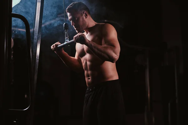 Fit sportsman concentrated on an excersise on a hand pull machine, pumping up his triceps in a dark gym surrounded by smoke — Stock fotografie