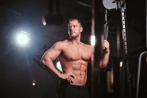 S'adapter athlète posant dans une salle de gym sombre entouré de fumée tout en faisant des extraits sur une machine à tirer la main — Photo