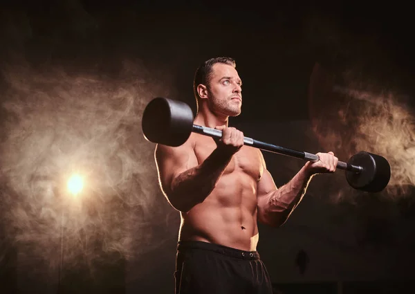 Shirtless bodybuilder doing weightlifts with barbell in a dark gym surrounded by smoke — 스톡 사진
