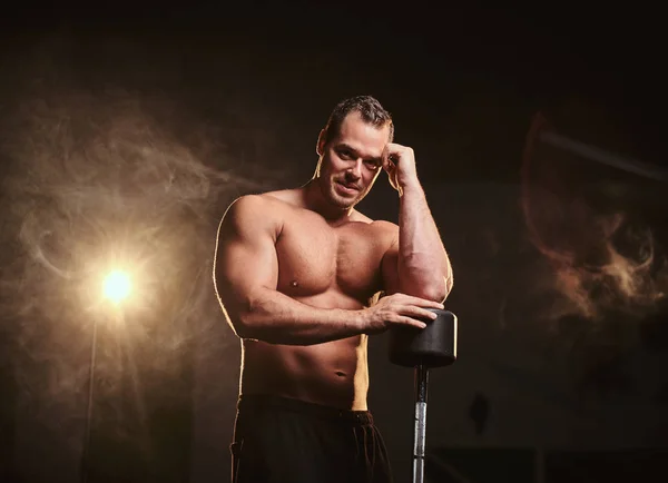 Athletic male leaning on the barbell in a thinking pose surrounded by smoke — Stock Photo, Image