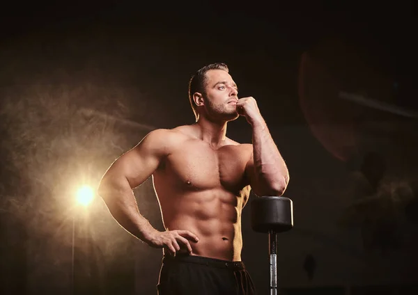 Athletic man with perfect body leaning on the barbell in a thinking pose in a dark gym surrounded by smoke — Stockfoto