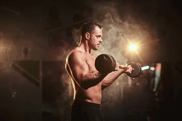 Shirtless bodybuilder doing weightlifts with barbell in a dark gym surrounded by smoke — 图库照片