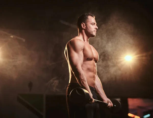 Shirtless bodybuilder doing weightlifts with barbell in a dark gym surrounded by smoke — Stockfoto