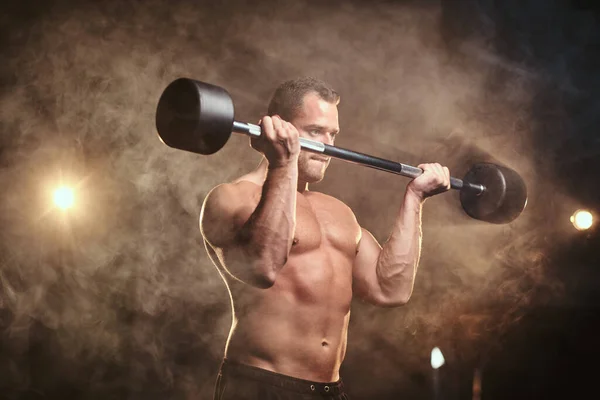 Shirtless bodybuilder doing weightlifts with barbell in a dark gym surrounded by smoke — Stock fotografie