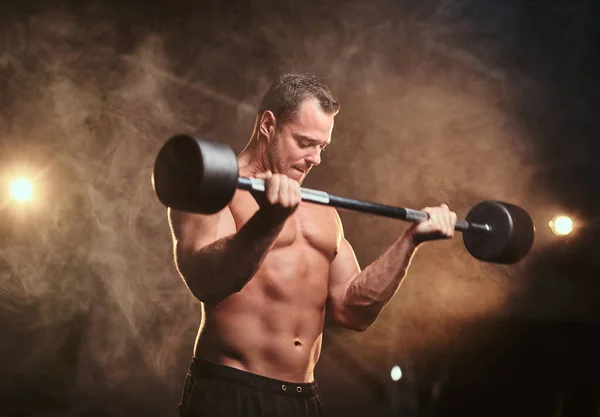 Shirtless bodybuilder doing weightlifts with barbell in a dark gym surrounded by smoke — 스톡 사진