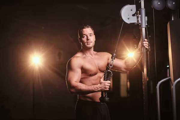 Fit sportsman concentrated on an excersise on a hand pull machine, pumping up his triceps in a dark gym surrounded by smoke — Stockfoto