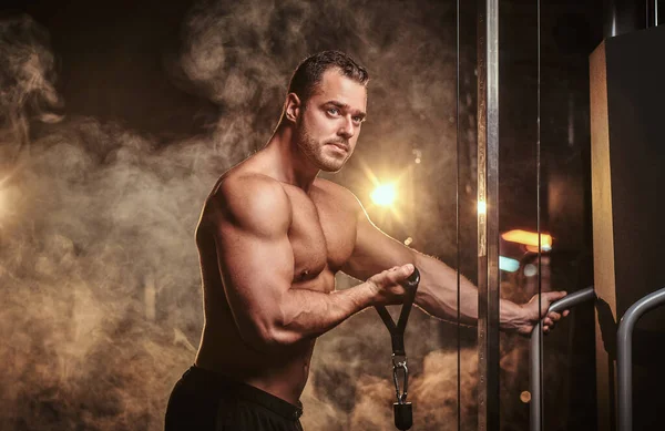 Fit athlete pumping up his biceps on a hand pull machine in a dark gym under the spotlights surrounded by smoke — Stockfoto