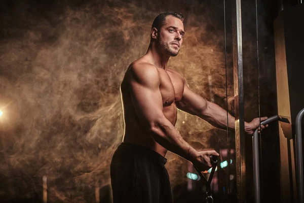 Fit athlete pumping up his biceps on a hand pull machine in a dark gym under the spotlights surrounded by smoke — Stockfoto