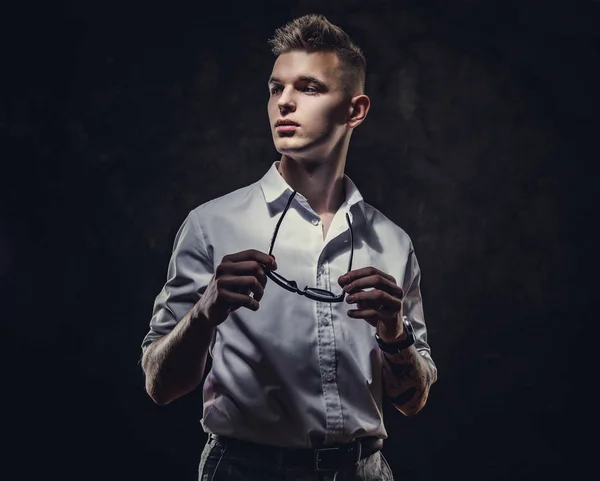 Bossy looking young adult male posing in a studio while holding his sunglasses in hands, isolated on a grey background — Stockfoto