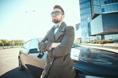 Genç, yakışıklı, yetişkin bir işadamı arabasının yanında dikiliyor, portreyi kapatıyor.