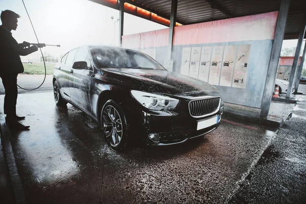 Adult man washing his car outdoors in a carwash under high pressured water