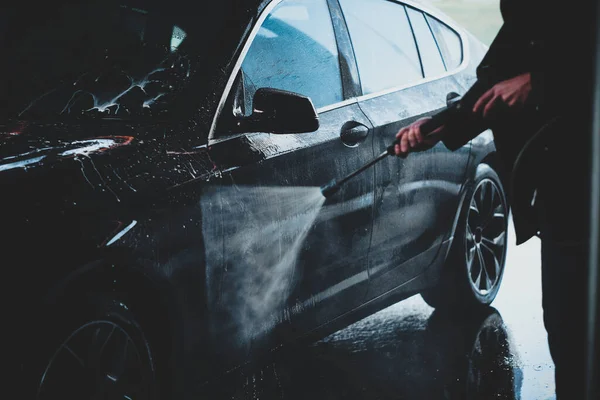 Yetişkin bir adam yüksek basınçlı suyun altında arabasını dışarıda yıkıyor. — Stok fotoğraf