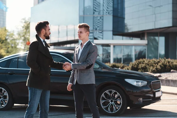 Двоє красивих бізнесменів тремтять руками перед чорним автомобілем, стоячи на відкритому повітрі — стокове фото