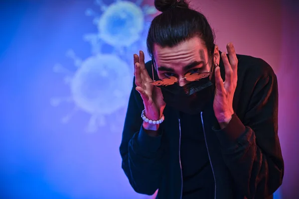 Stijlvolle jonge volwassen man draagt een medisch masker en ziet er bang uit in een neon studio. Covid-19-thema — Stockfoto
