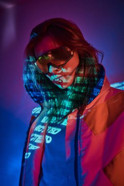 Tattooed racer girl standing over neon vivid text projection in a bright studio holding her glasses, close up look clipart