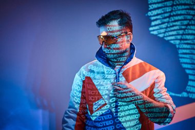 Tattoed racer man standing over neon vivid text projection in a bright studio showing his glasses clipart