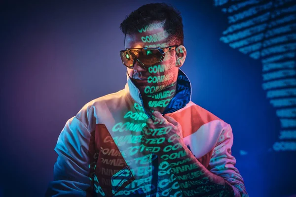 Tätowierte Rennfahrer männliches Modell steht über neon lebendige Textprojektion in einem hellen Studio hält ihre Brille, Nahaufnahme Blick — Stockfoto