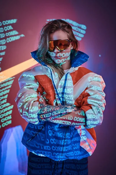 Tatuado piloto menina de pé sobre neon projeção de texto vívido em um estúdio brilhante segurando seus óculos, olhar de perto — Fotografia de Stock