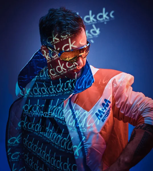 Tätowierter Racer-Mann steht über neonfarbener Textprojektion in einem hellen Studio und hält ihre Brille, Großaufnahme — Stockfoto