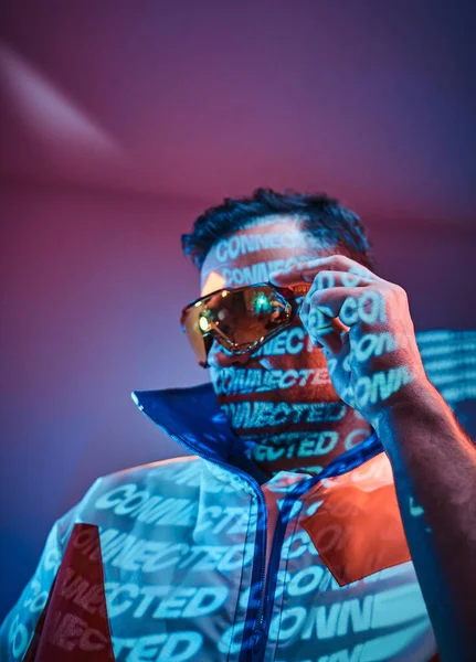 Homme racer tatoué debout sur néon projection de texte vif dans un studio lumineux portant des lunettes de soleil et manteau sportif — Photo