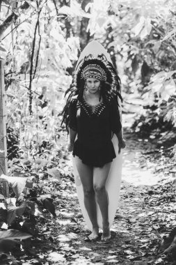 Ormanda sörf tahtasıyla poz veren çekici bir kadın Hint başlığı takıyor. Siyah beyaz fotoğraf.