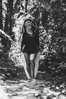 Ormanda sörf tahtasıyla poz veren çekici bir kadın Hint başlığı takıyor. Siyah beyaz fotoğraf.