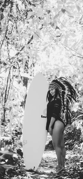 Attractive woman posing in forest with surfing board wearing mayan indian headwear. Black and white photo.