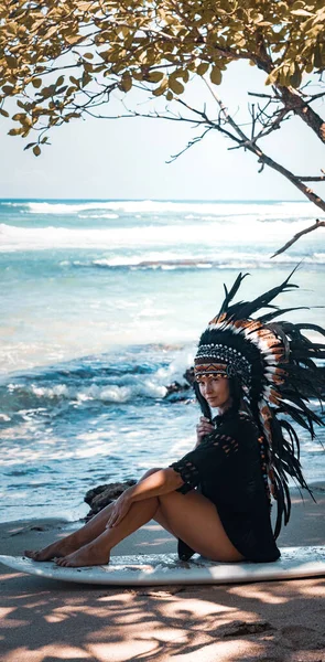 Attraktive und fitte Frau posiert auf einem Surfbrett und trägt Maya-Kopfbedeckungen an einem Strand in der Nähe der Meereswellen — Stockfoto