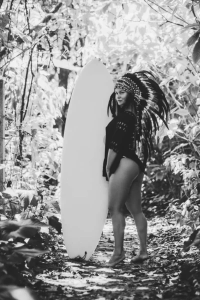 Aantrekkelijke vrouw die poseert in het bos met surfplank in Maya indiaanse hoofddeksels. Zwart-wit foto. — Stockfoto
