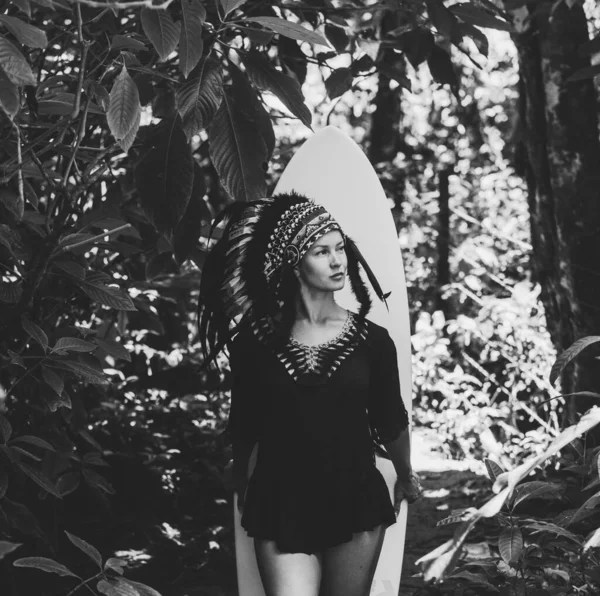 Mysterious and calm female model posing in a woods while holding a surfing board and wearing mayan feather hat